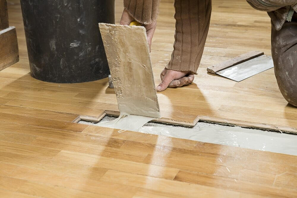 Close-up repair of oak strip Hardwood Floors in Eltham—installer trowelling adhesive to replace a tongue-and-groove section before sanding and refinishing.